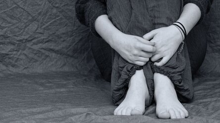 Teilansicht Person: diese sitzt mit angezogenen Knien und bloßen Füßen an eine Mauer gelehnt, die Hände umfassen die Beine