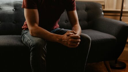 Mann sitzt aufrecht und mit mit gefalteten Händen auf einer Couch