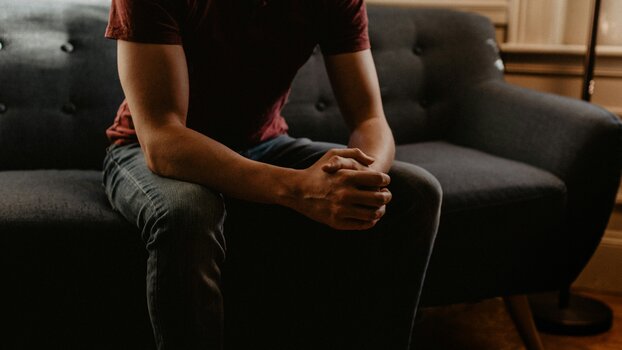 Mann sitzt aufrecht und mit mit gefalteten Händen auf einer Couch